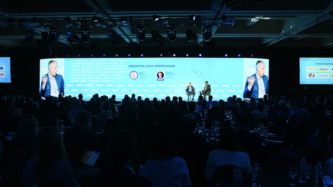 Auditorio colmado durante el 61° Coloquio IDEA con escenario iluminado y panel sobre competitividad bajo el lema “Juega Argentina”.