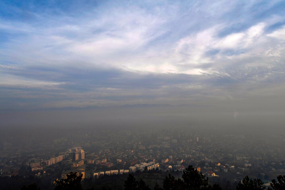 Vista panorámica de una ciudad europea cubierta por una capa de smog, con edificios y áreas urbanas parcialmente ocultas bajo una neblina densa y cielo nuboso.