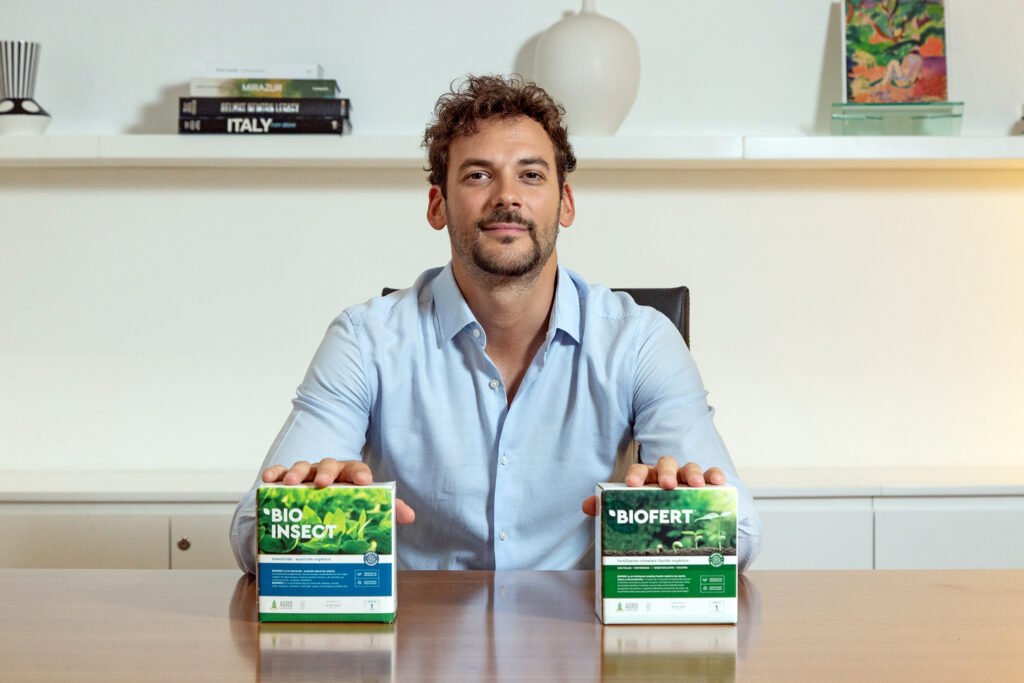 Joaquín Basanta sentado frente a una mesa sosteniendo dos productos bioinsumos de Agro Sustentable, Bio Insect y Biofert, en un entorno de oficina que refleja innovación agrícola y desarrollo tecnológico aplicado al campo.
